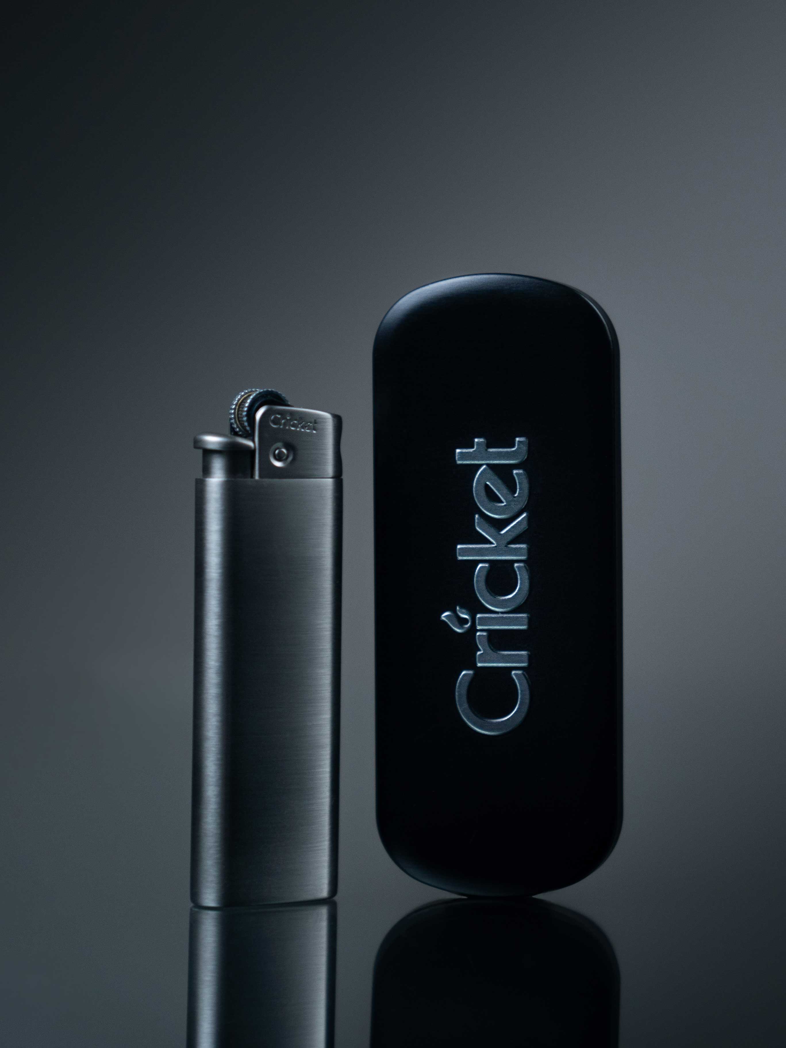 A silver Cricket metal lighter with a black branded tin case